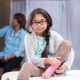 Girl wearing glasses sitting on an examining table and holding her ankle